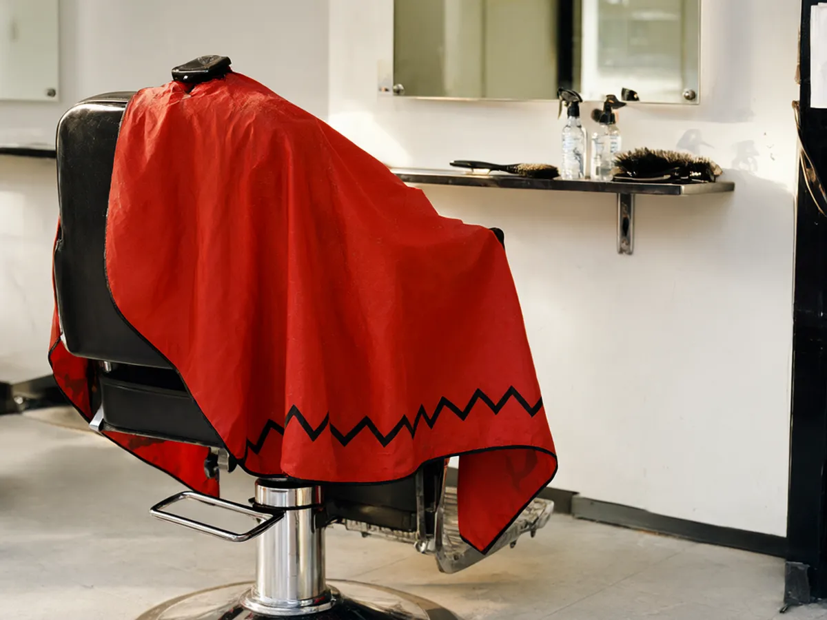 A black barber chair draped in a red cape with a black zigzag border in front of a mirror and a shelf of spray bottles